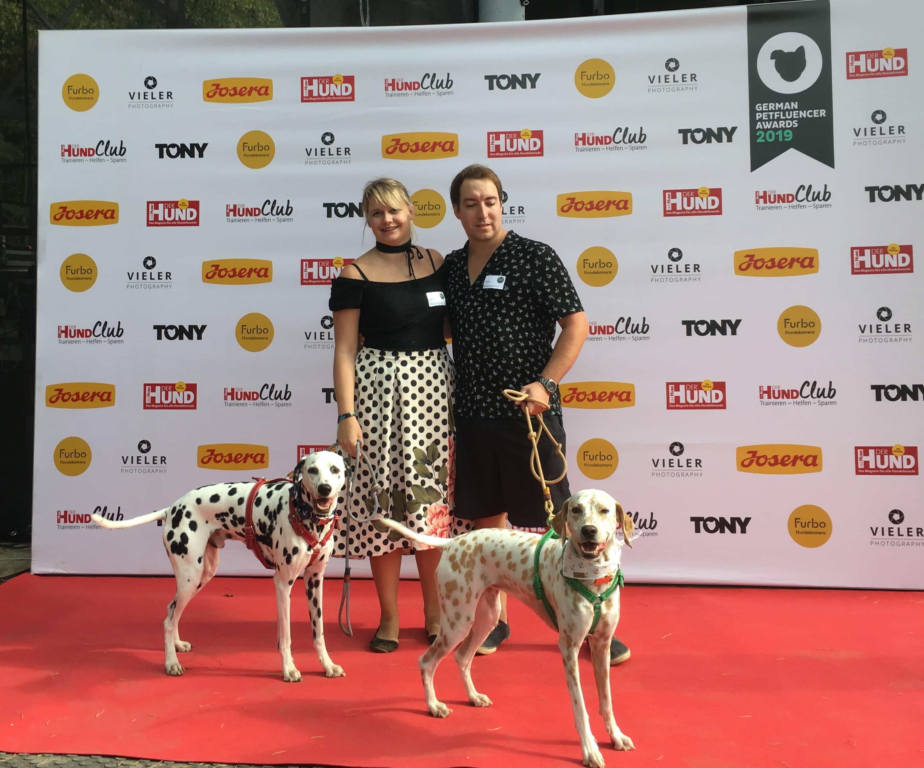 Zwei Personen mit zwei Dalmatinern posieren auf rotem Teppich vor Sponsorenwand