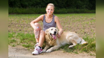 Frau hält eine Wasserflasche in der Hand und sitzt mit ihrem Golden Retriever am Wegrand, um eine Pause vom Training zu machen
