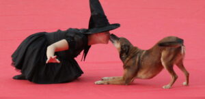 Frau im Hexenkostüm und Hund sitzen auf dem Boden, während der Hund in die Nase der Frau knabbert