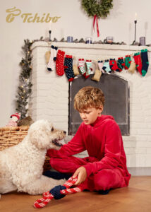 Ein Junge in einem roten Schlafanzug spielt auf dem Boden mit einem weißen Pudel. Hinter den beiden befindet sich ein weihnachtlich geschmückter Kamin.