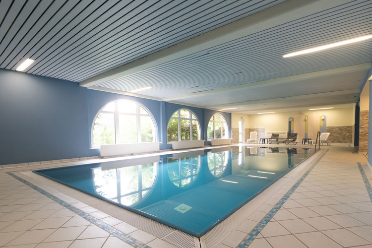 Helles, modernes Hallenbad mit großen Fenstern, Liegestühlen und blau gekacheltem Schwimmbecken im Vordergrund.