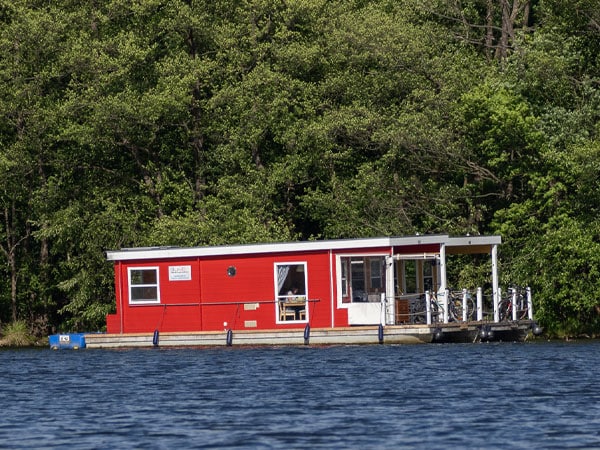Rotes Hausboot liegt am Ufer eines ruhigen Sees, umgeben von dichtem grünen Wald.