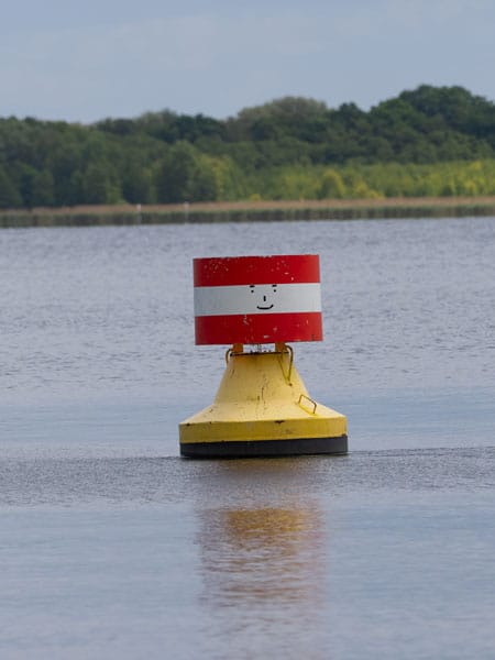 Gelbe Boje mit rot-weißem Aufsatz und Smiley-Gesicht schwimmt auf ruhigem Wasser vor Wald.