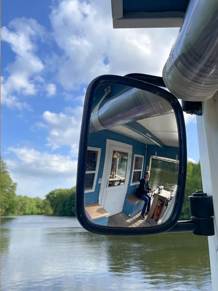 Spiegelung eines blauen Hausboots mit Person am Steuer auf ruhigem Fluss unter blauem Himmel. Spiegelung eines blauen Hausboots mit Person am Steuer auf ruhigem Fluss unter blauem Himmel.