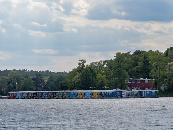 Bunte Hausboote liegen nebeneinander am Ufer, umgeben von Bäumen, unter bewölktem Himmel am See.