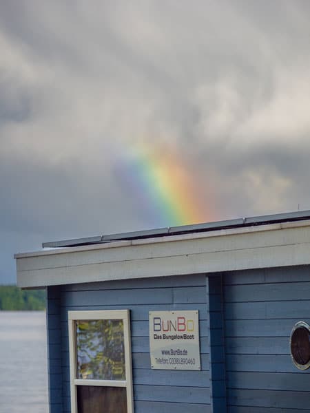 Blaues Hausboot am Wasser, über dem Dach wölbt sich ein Regenbogen vor wolkigem Himmel.