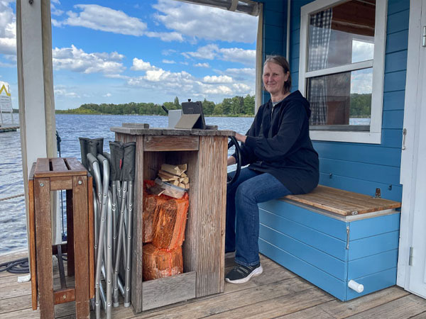 Person sitzt am Steuer eines blauen Hausboots am See, neben Holzvorrat und Klappstühlen.