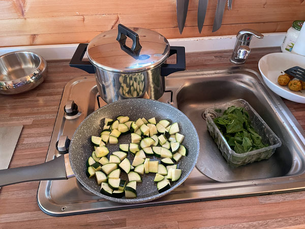 In der Küche stehen eine Pfanne mit Zucchini und ein Topf auf dem Herd, daneben frischer Spinat in der Spüle.