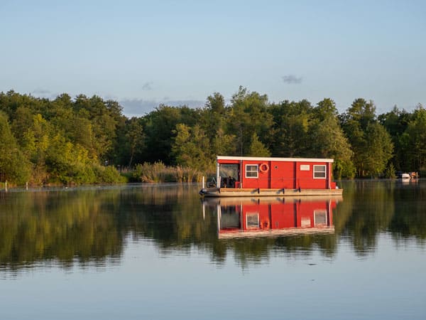 Rotes Hausboot spiegelt sich im ruhigen Wasser, dahinter dichter grüner Wald und blauer Himmel.