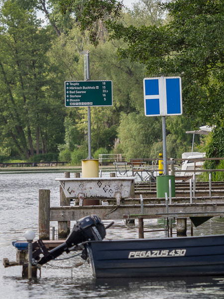 Steg mit Wegweisern und Boot am Ufer, umgeben von grünen Bäumen an einem ruhigen Gewässer.
