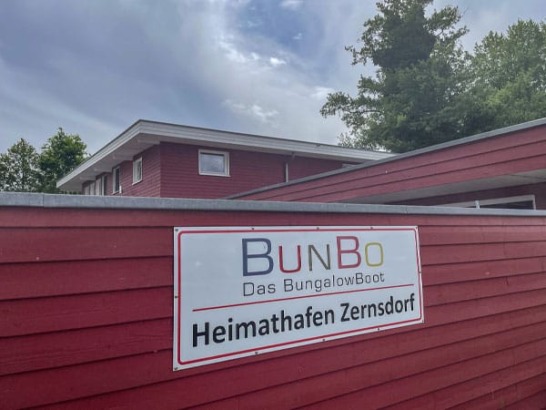 Rotes Gebäude mit Schild „BunBo Das BungalowBoot – Heimathafen Zernsdorf“ an einer Holzfassade.
