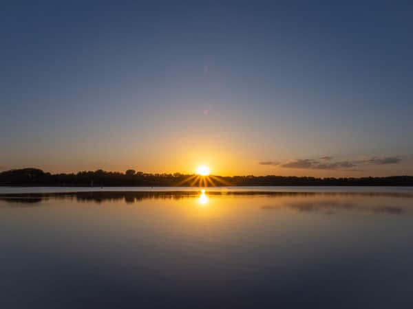 Sonnenaufgang spiegelt sich auf ruhigem See, klarer Himmel und dunkle Baumlinie am Horizont.