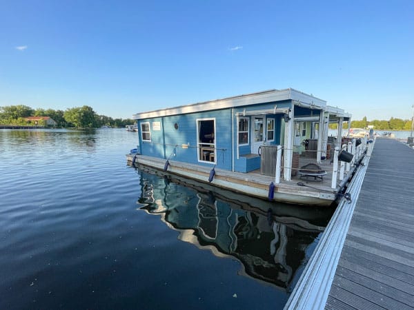Blaues Hausboot liegt an einem Steg auf ruhigem Wasser, umgeben von Bäumen und blauem Himmel.
