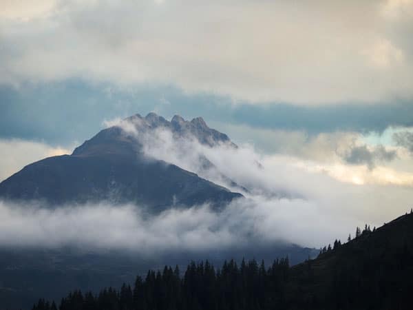 Berggipfel ragt mystisch aus tief liegenden Wolken, umrahmt von dunklen Tannen im Vordergrund.