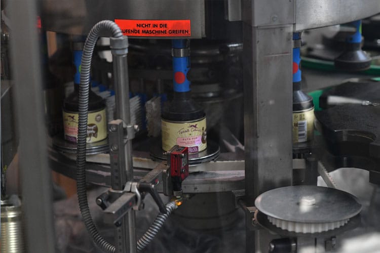 Dosenfutter wird in einer automatisierten Maschine mit Terra Canis-Etiketten versehen, Warnschild sichtbar.