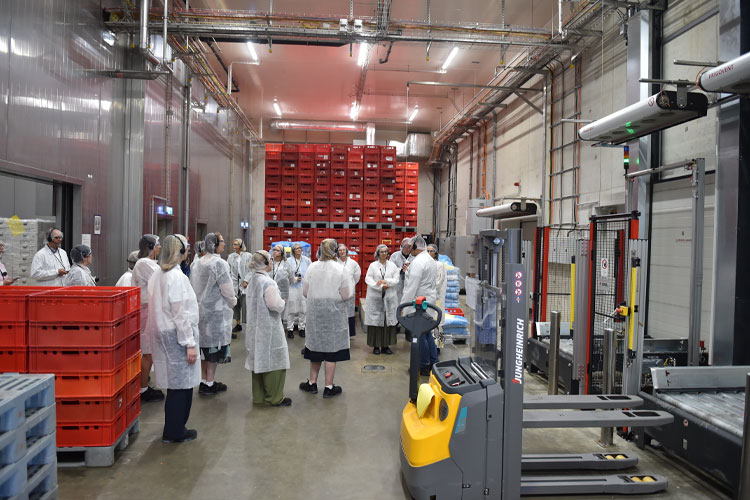Eine Besuchergruppe mit Schutzkleidung erhält eine Führung durch eine moderne Produktionshalle mit Maschinen und Kisten.