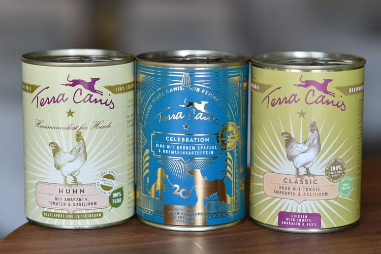 Drei Dosen Terra Canis Hundefutter (drei Sorten) stehen nebeneinander auf einem Holztisch, in der Mitte die Jubiläums-Edition