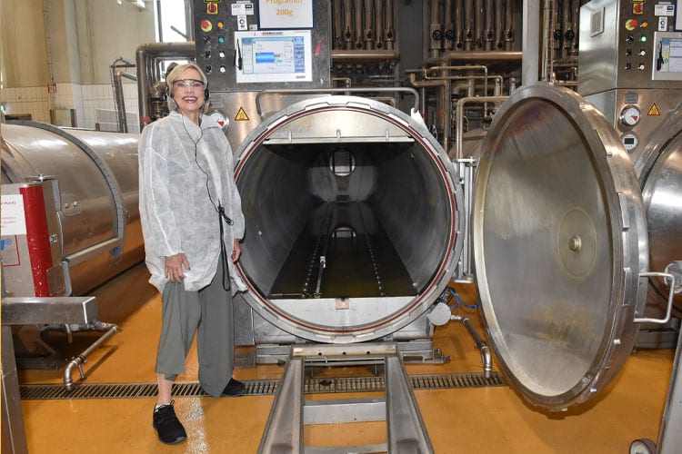 Nina Ruge in Schutzkleidung steht neben großem, geöffneten Edelstahl-Autoklaven in einer Produktionshalle.