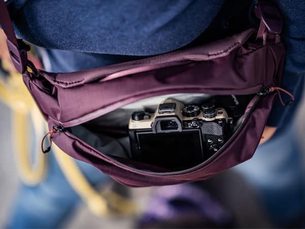 Kompakte OM SYSTEM Kamera liegt griffbereit in einer geöffneten, bordeauxroten Bauchtasche.