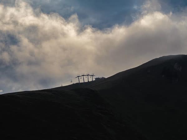 Dunkler Bergrücken mit Silhouette einer Seilbahn vor dramatisch bewölktem Himmel in den Alpen.
