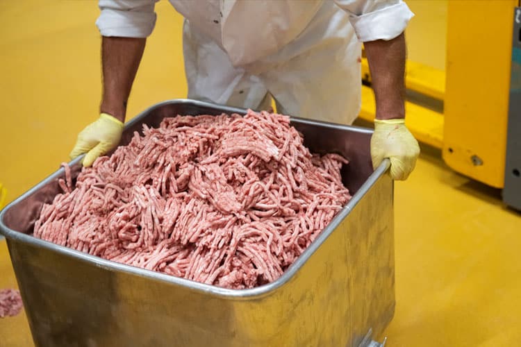 Person in weißem Kittel und Handschuhen schiebt Wanne voll frischem zerkleinertem Fleisch.
