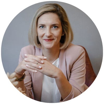 Frau mit blonder Bobfrisur und rosa Blazer sitzt entspannt an einem Tisch.