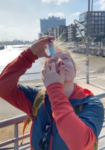 Person verwendet Augentropfen am Hamburger Hafen vor der Elbphilharmonie bei sonnigem Wetter.
