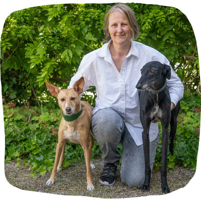 Susanne C. Steiger in der Hocke vor grünem Hintergrund, mit den Armen um die Hündinnen Paula und Lora.