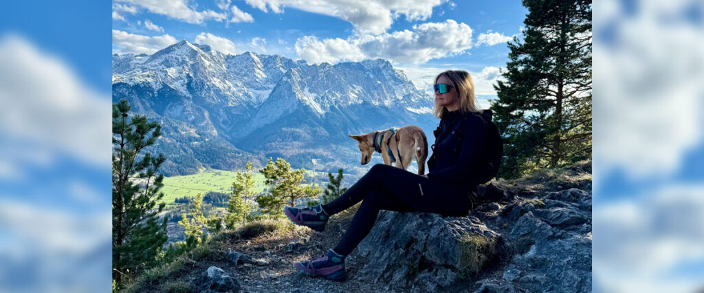 Frau sitzt mit Hund auf Felsen vor beeindruckender Alpenkulisse unter blauem Himmel mit Wolken.