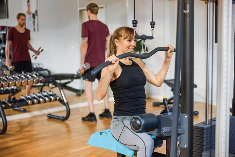Eine Frau trainiert an einer Kraftmaschine im Fitnessstudio, im Hintergrund trainieren weitere Personen.