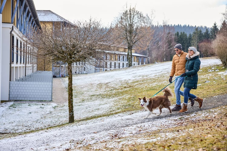 Ein Paar spaziert mit Hund an der Leine auf einem verschneiten Weg vor dem Club Aldiana im Grünen.