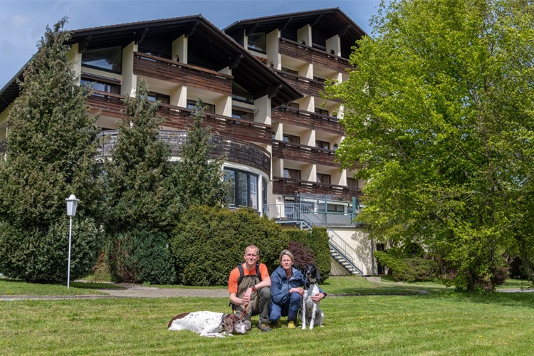 Zwei Personen mit zwei Hunden auf einer Wiese vor dem großen Wolfshof Hotelgebäude, umgeben von Bäumen.