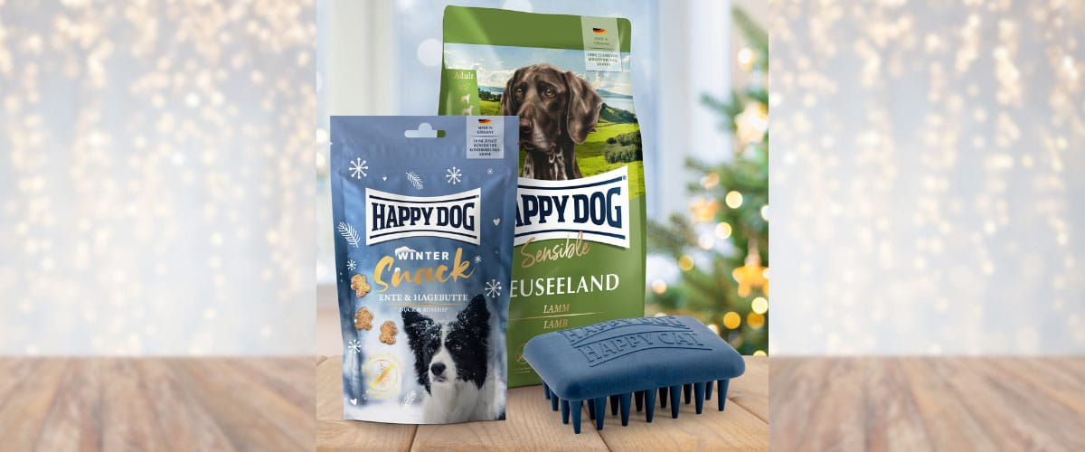 Happy Dog Winter Snack, Trockenfutter Neuseeland und eine blaue Bürste stehen vor festlichem Hintergrund auf Holz.