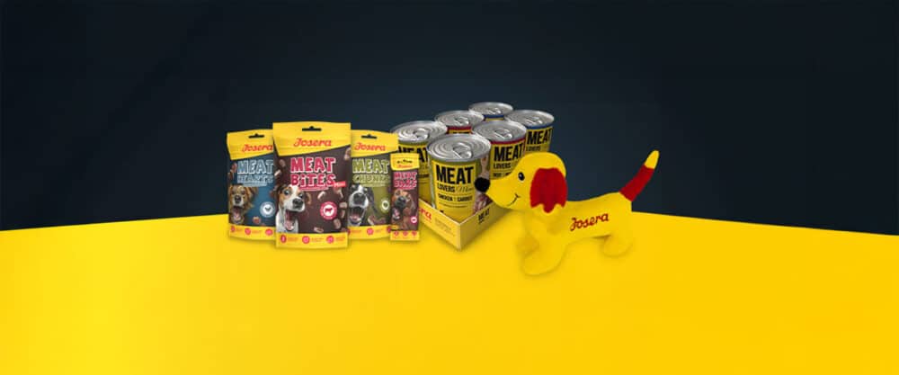 Josera Hundefutter und Snacks als Tüten und Dosen sind neben einem gelben Stoffhund auf gelb-schwarzem Hintergrund angeordnet.