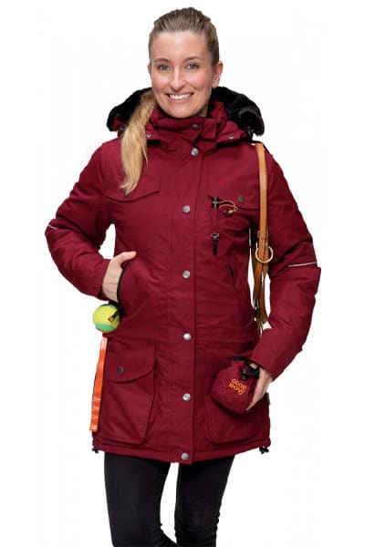 Frau trägt eine dunkelrote Winterjacke mit Kapuze und Hundezubehör, darunter Leckerlibeutel und Ball.