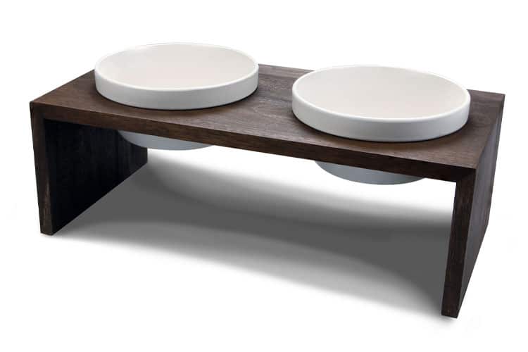 Zwei weiße Futternäpfe stehen in einer erhöhten, dunklen Holzhalterung auf weißem Hintergrund.