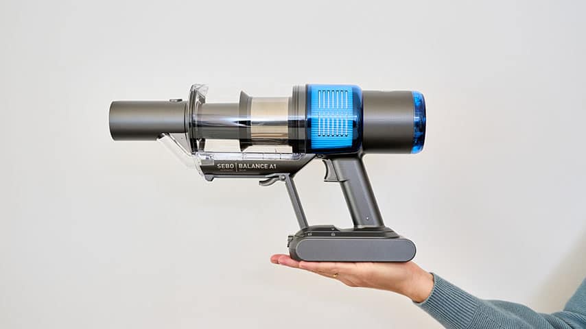 Eine Person hält einen kompakten, modernen Akkustaubsauger mit blauem Filteraufsatz vor neutralem Hintergrund.
