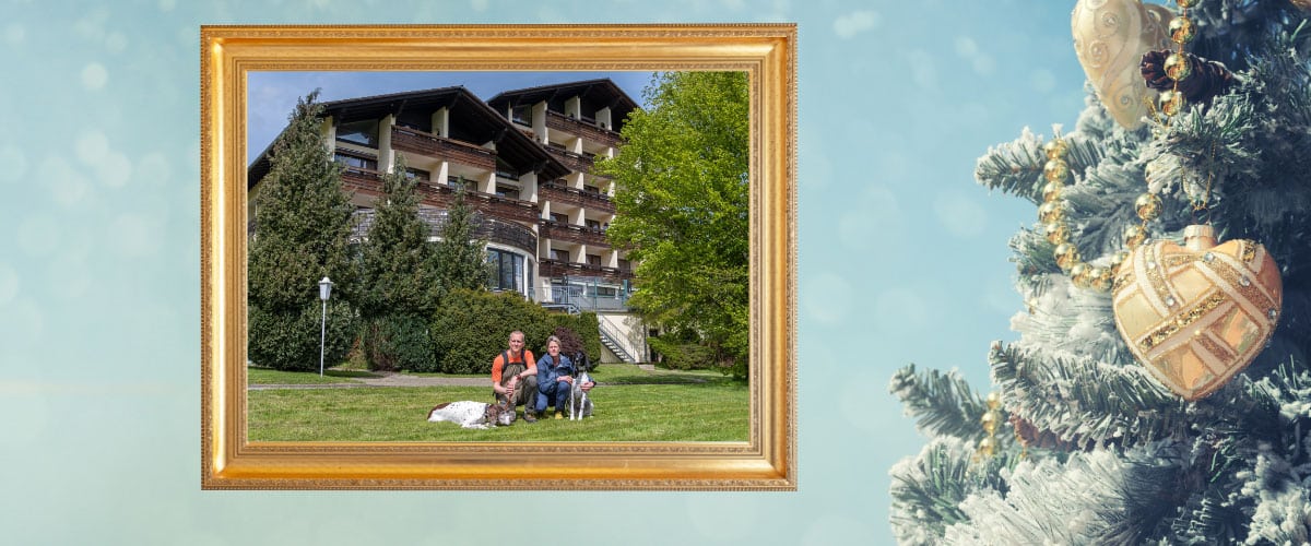Gold gerahmtes Foto von zwei Menschen und zwei Hunden vor dem Wolfshof Hotelgebäude, daneben ein geschmückter Weihnachtsbaum.