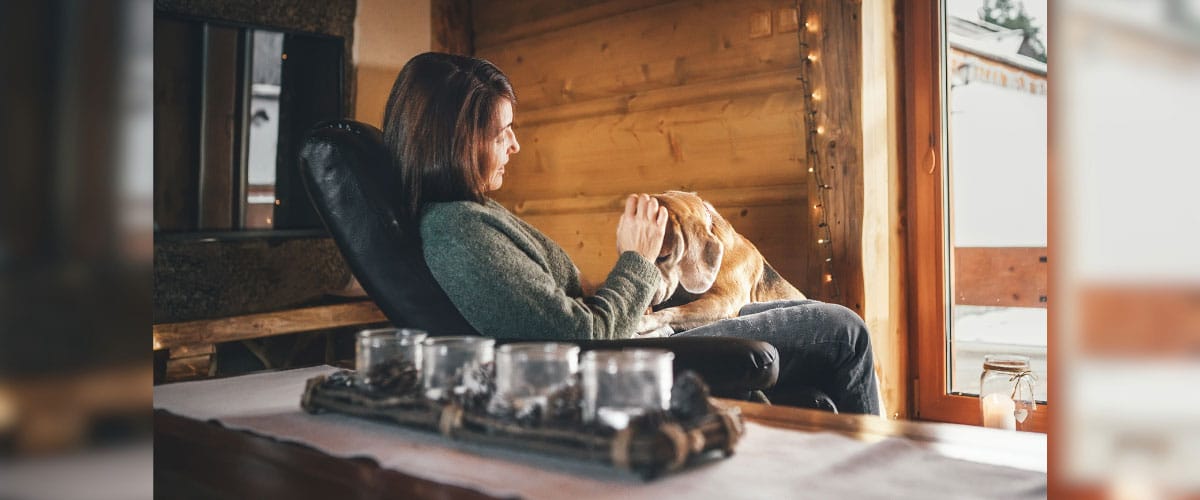 Frau sitzt entspannt im Sessel am Fenster und streichelt ihren Hund in gemütlichem Hotelzimmer.