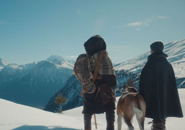 Zwei Personen mit Hund gehen auf Weg durch verschneite Berge in winterlicher Landschaft.