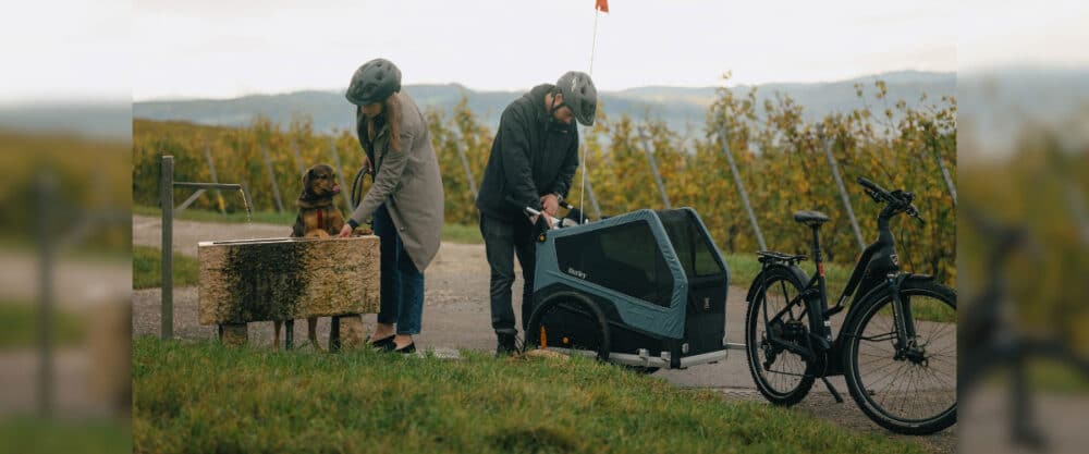 Zwei Personen mit Fahrrad und Hundeanhänger machen Pause, während ein Hund Wasser trinkt.