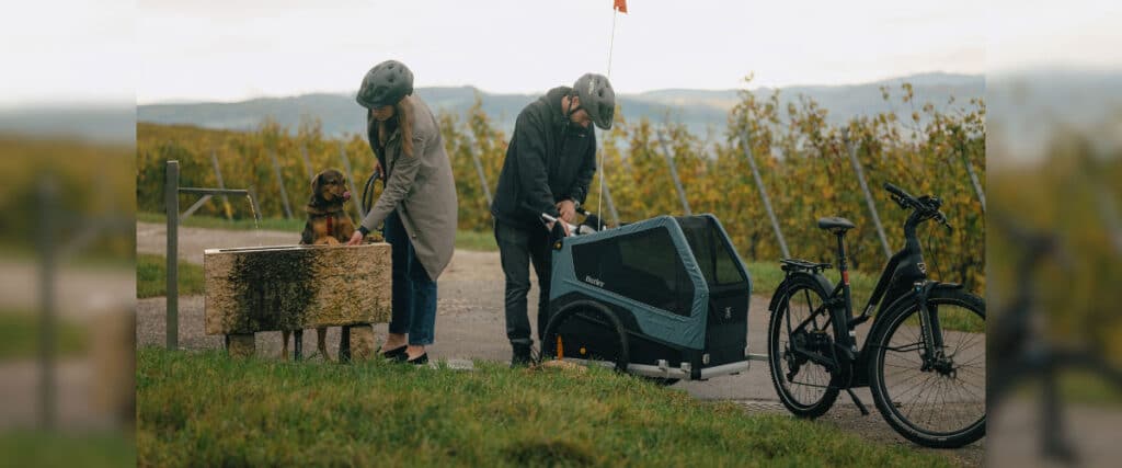 Zwei Personen mit Fahrrad und Hundeanhänger machen Pause, während ein Hund Wasser trinkt.