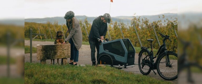 Zwei Personen mit Fahrrad und Hundeanhänger machen Pause, während ein Hund Wasser trinkt.