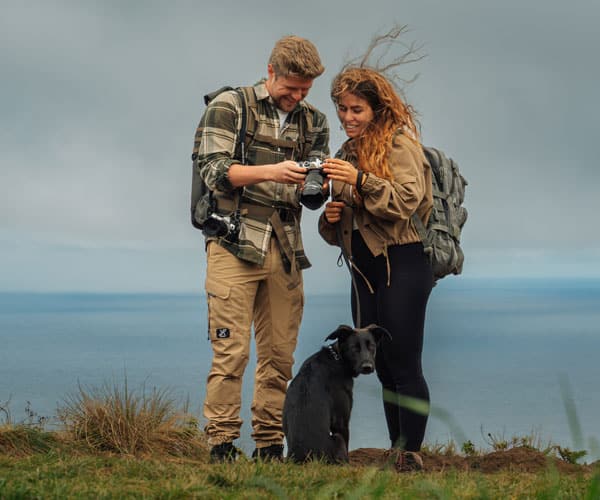 Mann und Frau mit Kamera betrachten Fotos am Meereskliff, ein Hund sitzt zwischen ihnen.