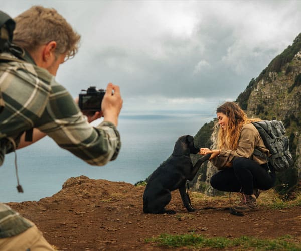 Mann fotografiert Hund, der auf Aussichtspunkt am Meer hockender Frau die Pfote gibt.