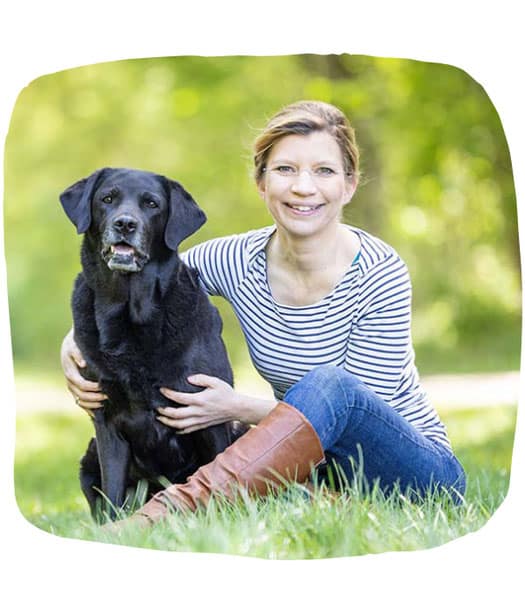 Kristina Ziemer-Falke in Jeans, braunen Stiefeln und gestreiftem Oberteil sitzt neben einem schwarzen Hund auf einer Wiese.