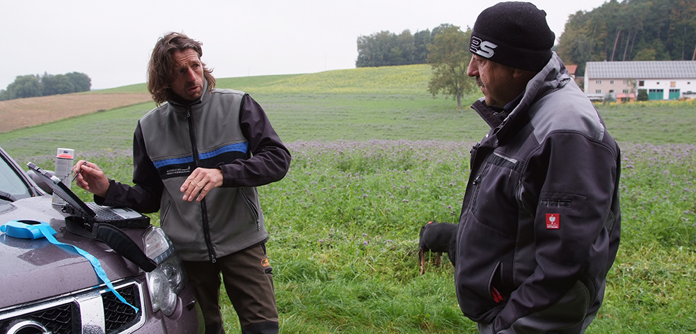 Förster Brandl spricht mit Waldbesitzer an Geländewagen mit Laptop, im Hintergrund Feld und Hund im Gras.