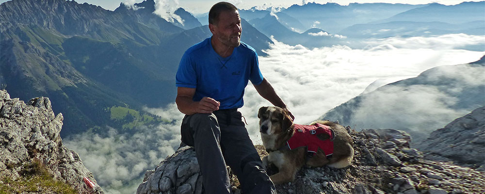 Wanderer sitzt mit Hund im roten Geschirr auf Felsen, umgeben von Alpenpanorama und Wolkenmeer im Tal.