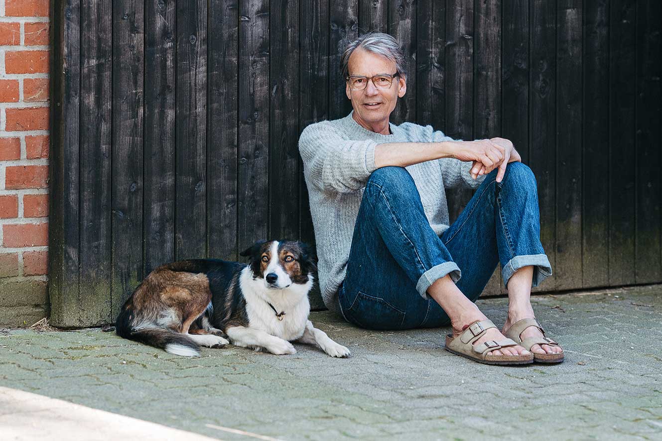 Michael Grewe sitzt auf dem Boden vor einem Scheunentor. Rechts daneben liegt ein Hund.